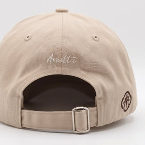 Close-up rear view of a beige baseball cap with 'Anatta' embroidered in white script on the back, a dark brown side logo, and an adjustable metal buckle strap.