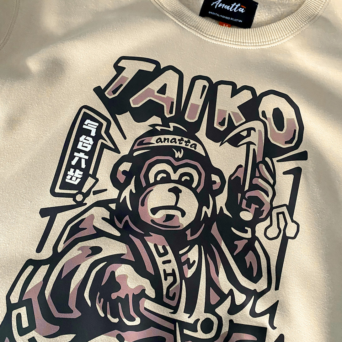 Taiko - a close-up of the design of a monkey dressed in traditional Japanese clothing playing a taiko drum, printed on the front of a khaki sweatshirt -1 