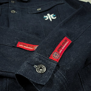 Close-up of the Rogue Wave denim jacket showing red woven sleeve labels, metal buttons, chest embroidery, and textured indigo denim.