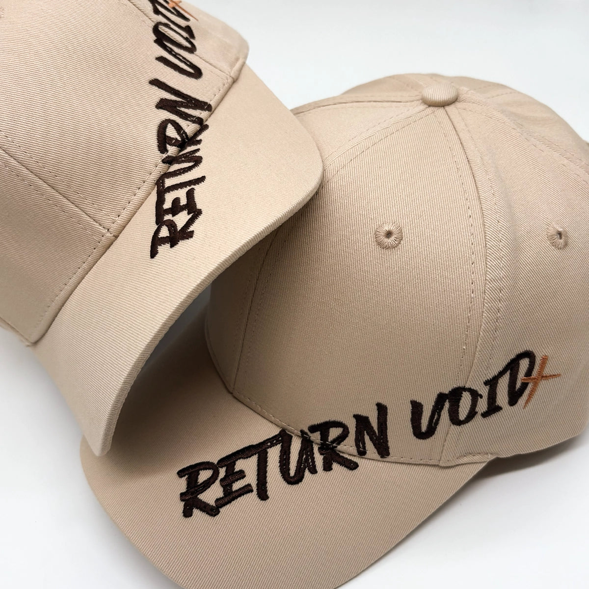 Two beige baseball caps with embroidered text reading 'RETURN VOID+' on the brim and side, shown on a clean white background.