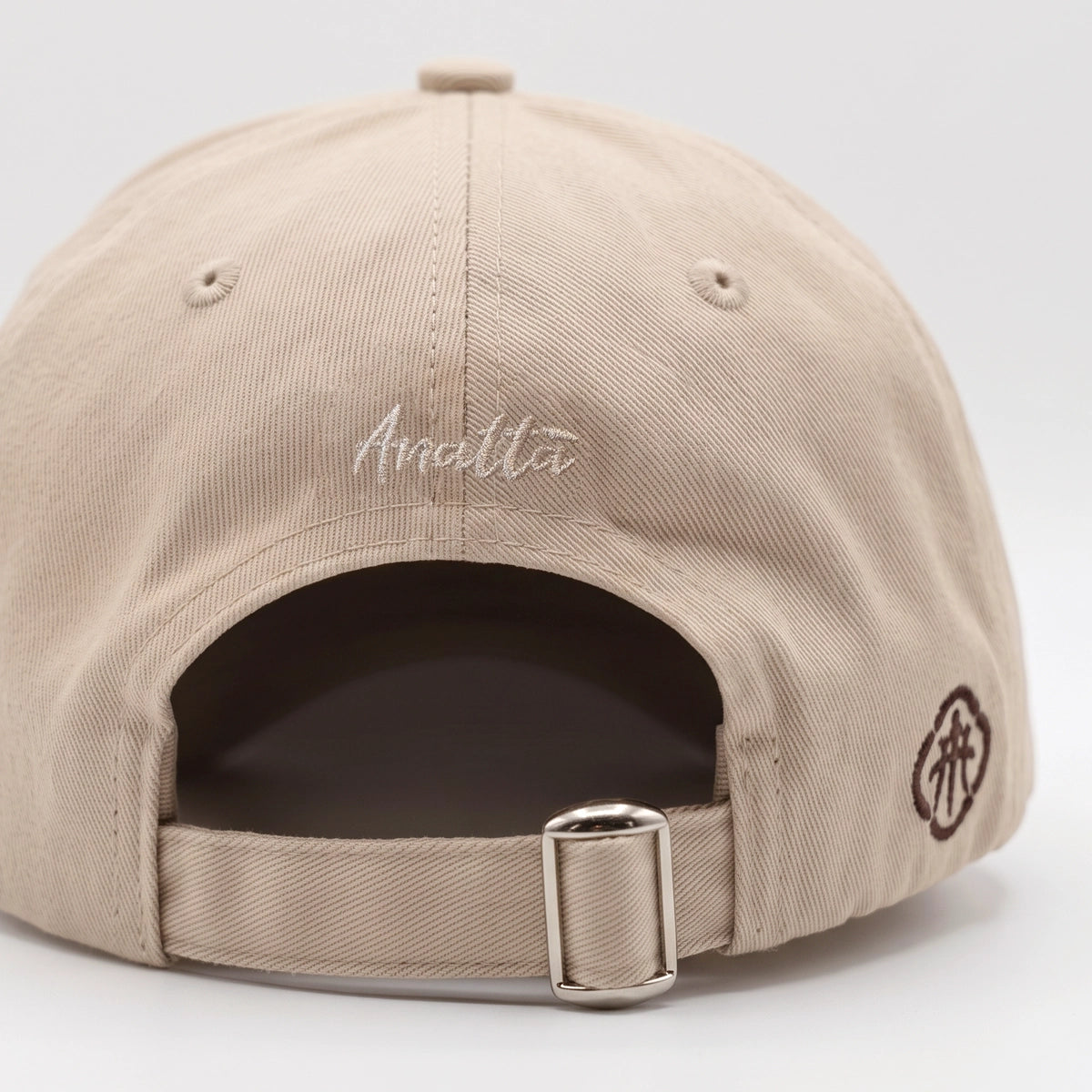 Close-up rear view of a beige baseball cap with 'Anatta' embroidered in white script on the back, a dark brown side logo, and an adjustable metal buckle strap.