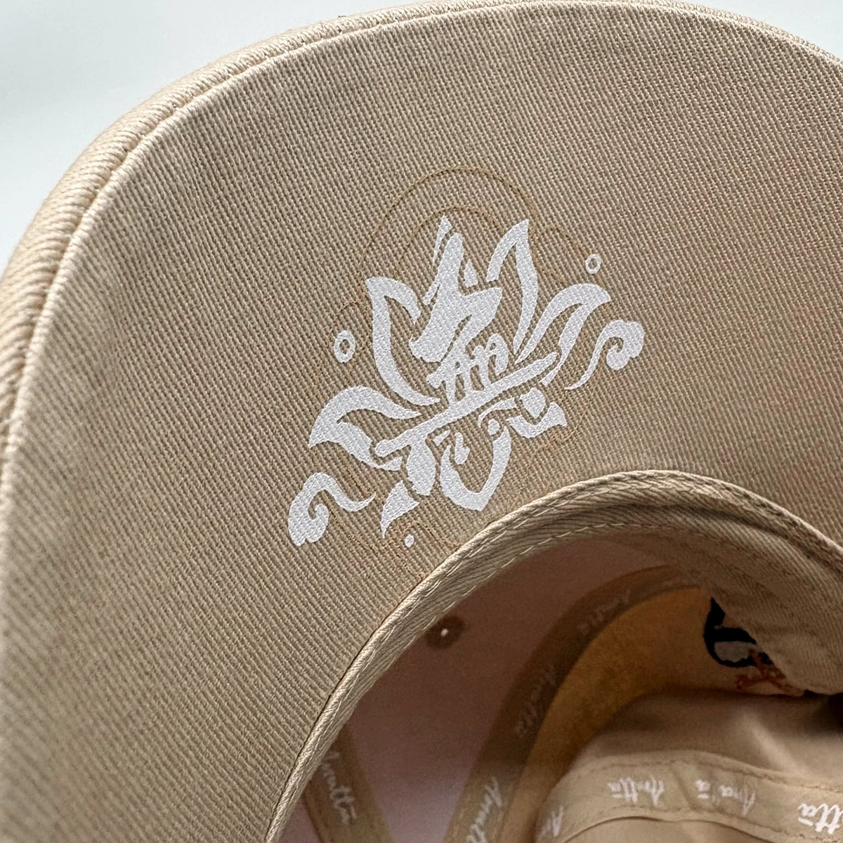 Close-up of a beige baseball cap showing the curved brim with a white lotus-style graphic and the interior crown.