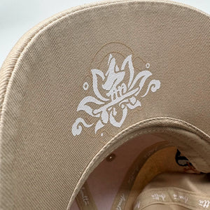 Close-up of a beige baseball cap showing the curved brim with a white lotus-style graphic and the interior crown.