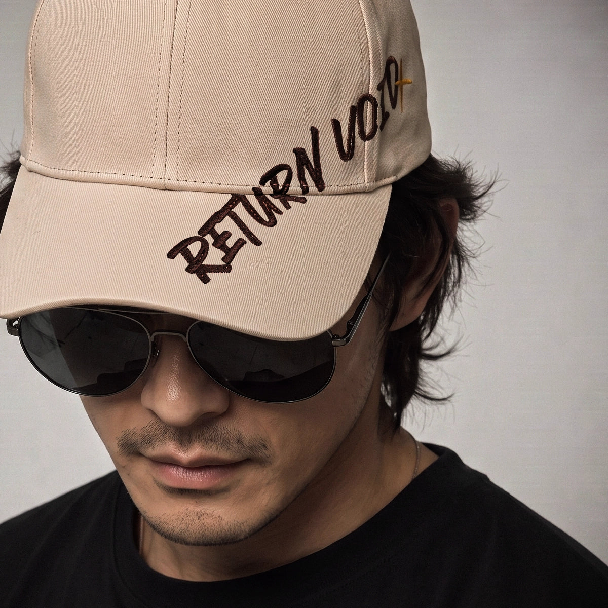 A man wearing a beige baseball cap with 'RETURN VOTE' embroidered on the brim, paired with dark sunglasses and a black shirt against a plain light gray background.