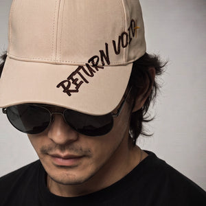 A man wearing a beige baseball cap with 'RETURN VOTE' embroidered on the brim, paired with dark sunglasses and a black shirt against a plain light gray background.