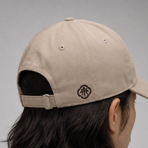 A close-up side view of a person wearing a beige baseball cap, showing embroidered logo, and side embroidery against a neutral gray background.
