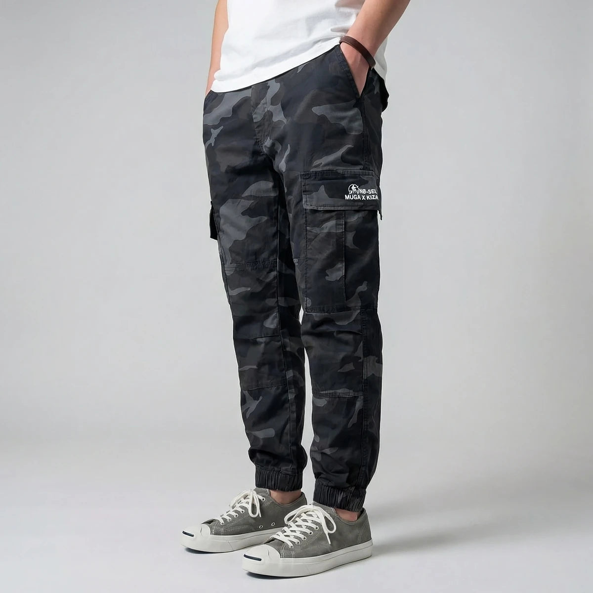 Model wearing black camouflage cargo pants with a visible brand logo on a gray background- Anatta Streetwear