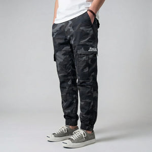 Model wearing black camouflage cargo pants with a visible brand logo on a gray background- Anatta Streetwear