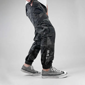 model wearing black camouflage pants with jogger fit on a gray background- Anatta Streetwear-2
