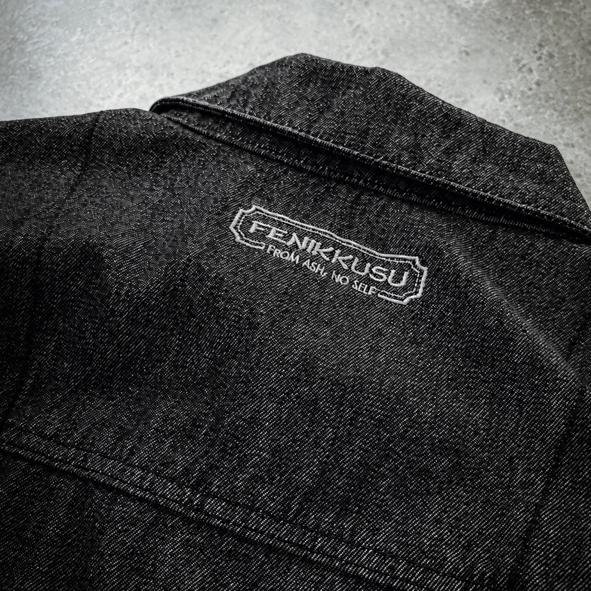 Close-up of the Fenikkusu embroidered back-neck logo on the dark grey Japanese denim jacket