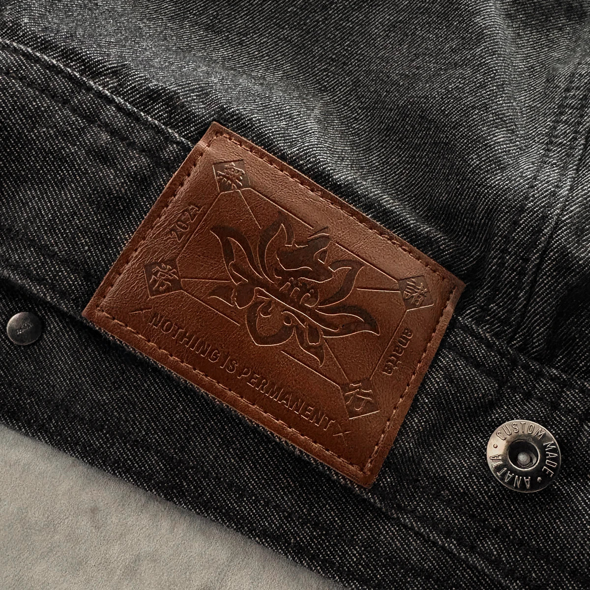 Close-up of the brown leather brand patch and metal hardware on the Fenikkusu dark grey Japanese denim jacket