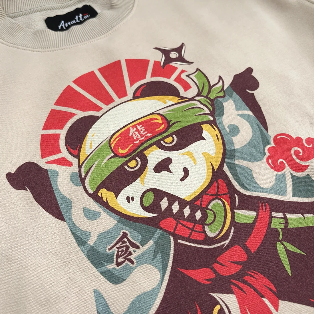 Close-up of a khaki sweatshirt featuring a colorful graphic of a panda in a samurai mask with a green headband, red cloud, and blue wave patterns.