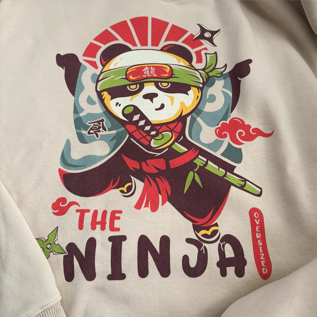 A close-up of a khaki sweatshirt featuring a colorful graphic of a panda dressed as a ninja with the text 'THE NINJA' and 'OVERSIZED'.