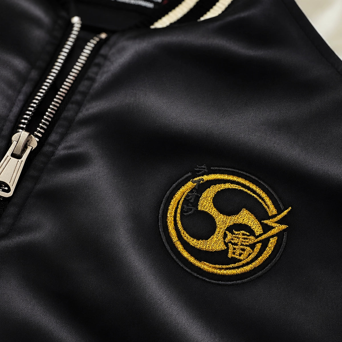 Close-up of the Raijin’s Wrath Japanese satin jacket, showing the gold embroidered chest logo and lustrous black satin fabric