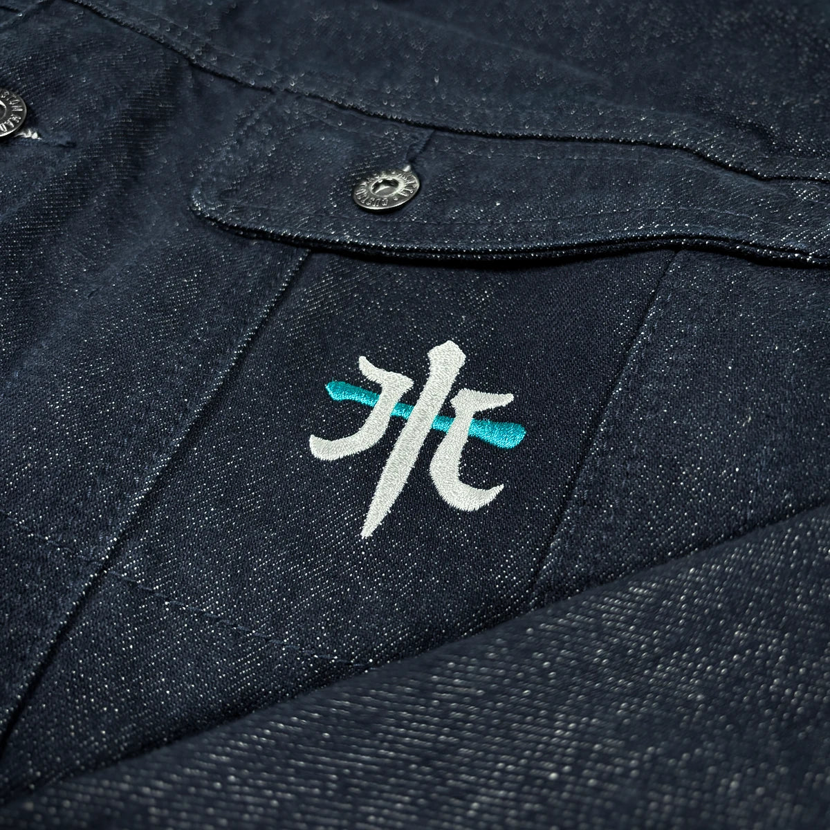 Close-up of the Rogue Wave denim jacket showing the embroidered chest symbol and textured indigo denim.