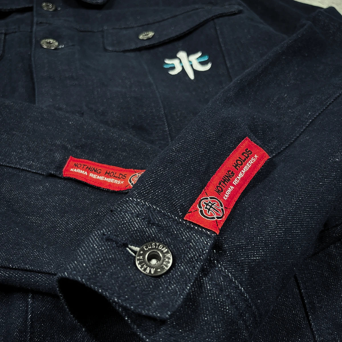 Close-up of the Rogue Wave denim jacket showing red woven sleeve labels, metal buttons, chest embroidery, and textured indigo denim.